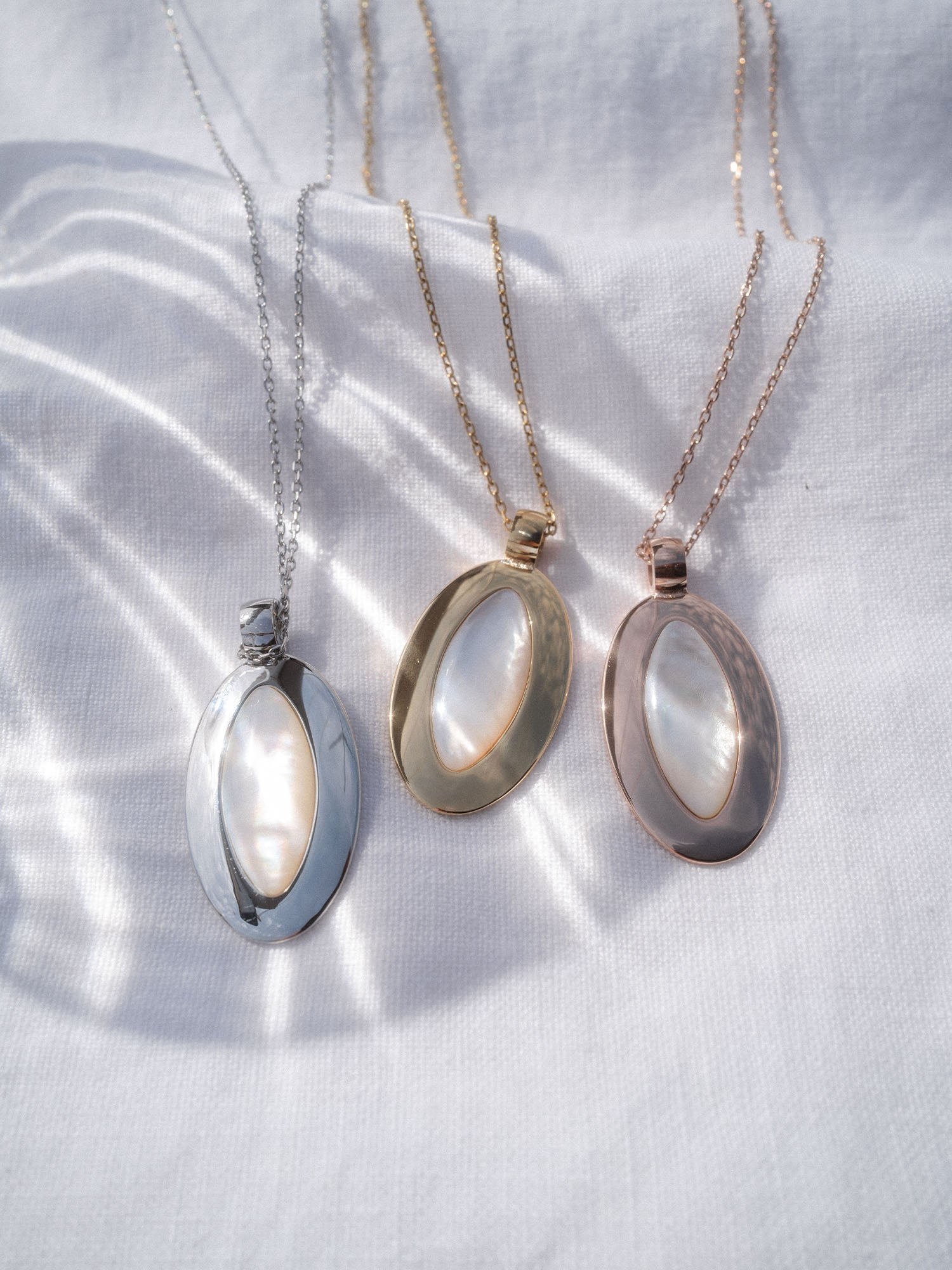 Island Elegance Kette von PURELEI mit ovalem Perlmutt-Anhänger in Silber, Gold und Roségold auf weißem Stoff.